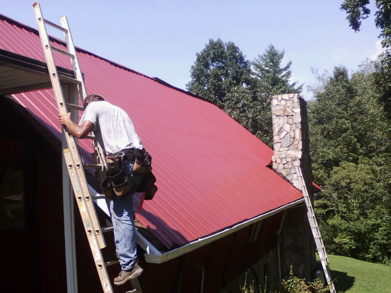 Roofer on a ladder beside a red metal roof and stone chimney — Soffit & Fascia Repair by Roofing Brothers in Palos Heights